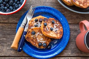 French toast on a blue plate on a picnic table, topped with coconut and blueberries